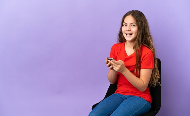 Petite fille caucasienne assise sur une chaise isolée sur fond violet surprise et envoyant un message