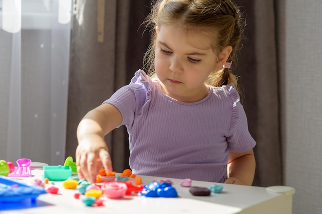 Petite fille caucasienne blonde jouant avec de la pâte à modeler multicolore sur une table blanche jouant avec de la pâte à modeler à la maison concept