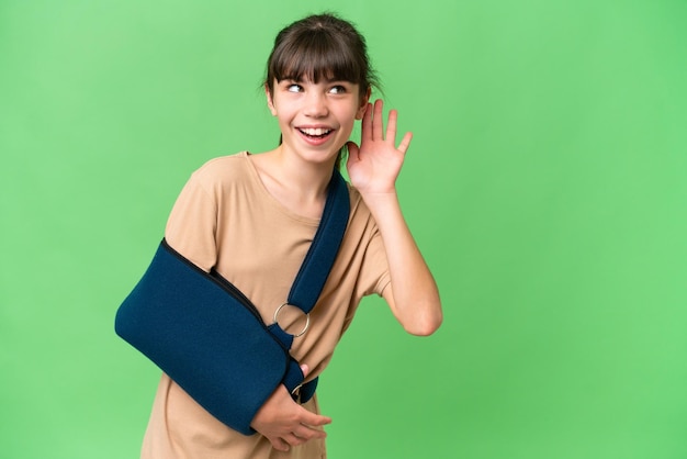 Petite fille caucasienne avec bras cassé et portant une écharpe sur fond isolé écoutant quelque chose en mettant la main sur l'oreille