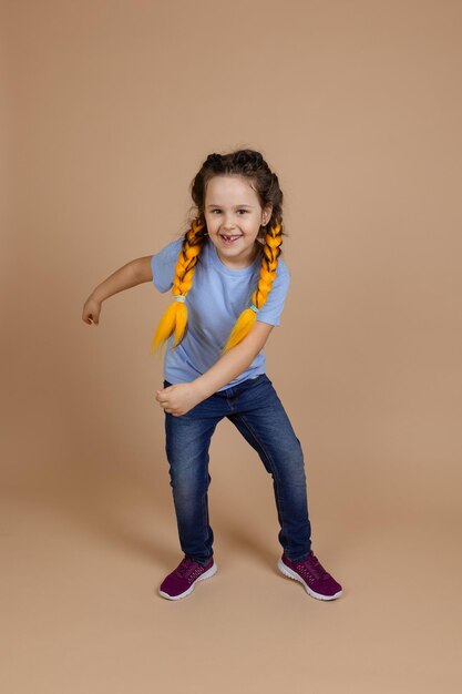 Petite fille caucasienne brillante avec des tresses kanekalon jaunes dansant les bras mobiles souriant avec une dent manquante regardant la caméra sur fond beige portant un t-shirt bleu et un jean