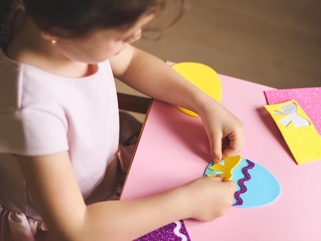 Une petite fille caucasienne colle un arc jaune sur un œuf en feutre bleu avec ses mains