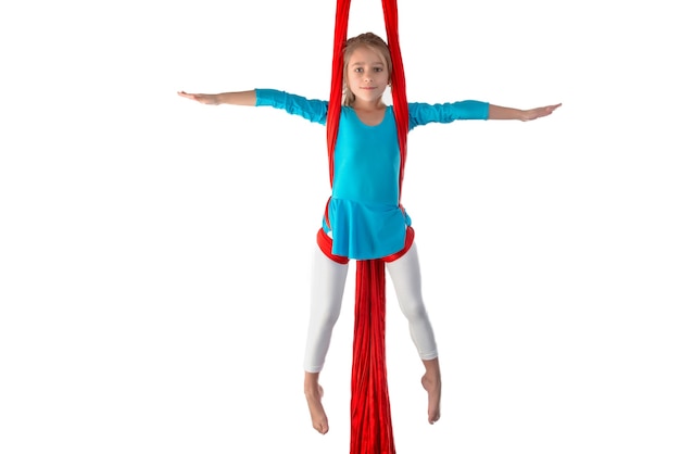 Petite fille caucasienne concentrée dans un maillot de bain bleu fait des exercices de gymnastique sur un ruban aérien rouge sur fond blanc. Flexibilité de la gymnastique pour les enfants. Espace publicitaire