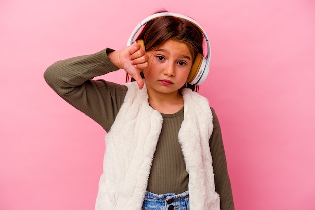 Petite fille caucasienne écoutant de la musique isolée sur fond rose montrant un geste d'aversion, les pouces vers le bas. Notion de désaccord.