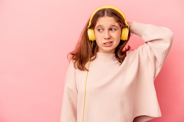 Petite fille caucasienne écoutant de la musique isolée sur fond rose touchant l'arrière de la tête, pensant et faisant un choix.