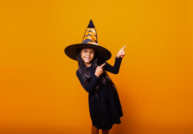 Petite fille caucasienne gaie dans un costume de sorcière d'Halloween pointe son doigt sur le côté.