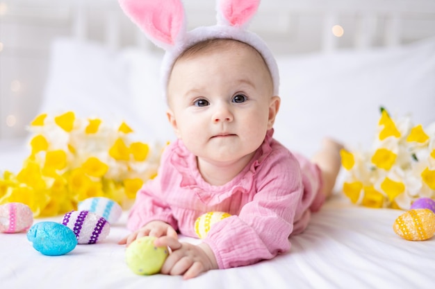 Une petite fille caucasienne heureuse avec un bord d'oreilles de lapin sur la tête se trouve sur le lit à la maison avec des oeufs de Pâques colorés et des fleurs de printemps jaunes regarde la caméra rit bébé de Pâques