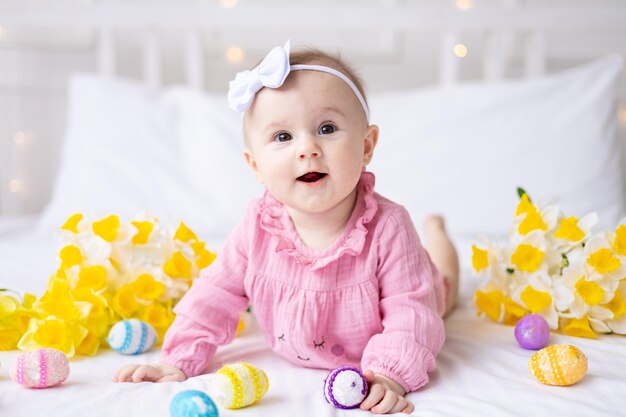 Une petite fille caucasienne heureuse avec un bord d'oreilles de lapin sur la tête se trouve sur le lit à la maison avec des oeufs de Pâques colorés et des fleurs de printemps jaunes regarde la caméra rit bébé de Pâques