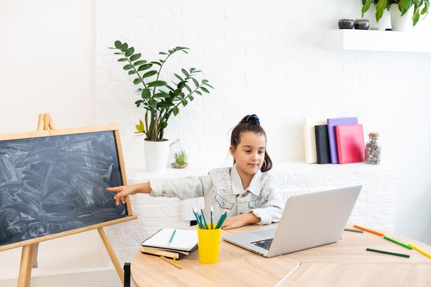 Une petite fille caucasienne intelligente s'assoit à la table et étudie sur un ordinateur portable, prend des notes dans un cahier, une petite écolière écrit à la main, prépare des devoirs, prépare une tâche à la maison, concept d'éducation à distance