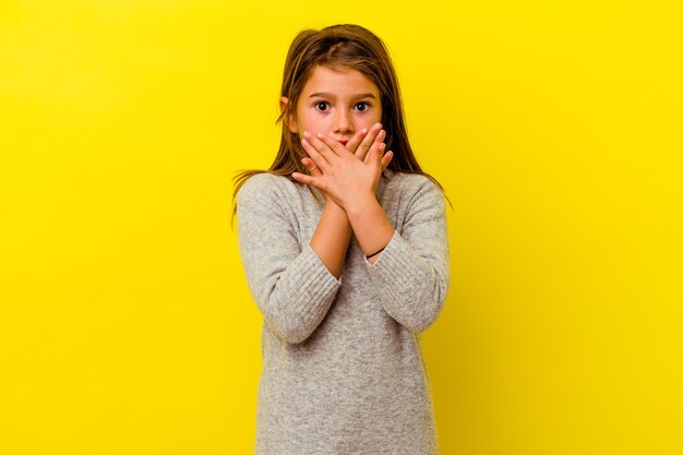 Petite fille caucasienne isolée sur bouche couvrant jaune choqué avec les mains.