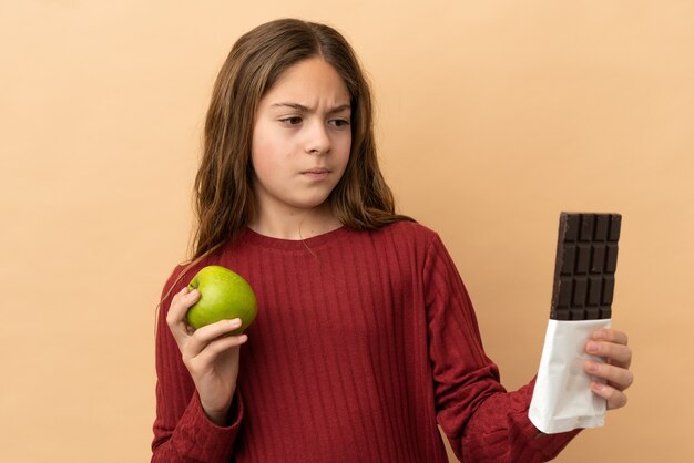 Petite fille caucasienne isolée sur fond beige ayant des doutes en prenant une tablette de chocolat dans une main et une pomme dans l'autre