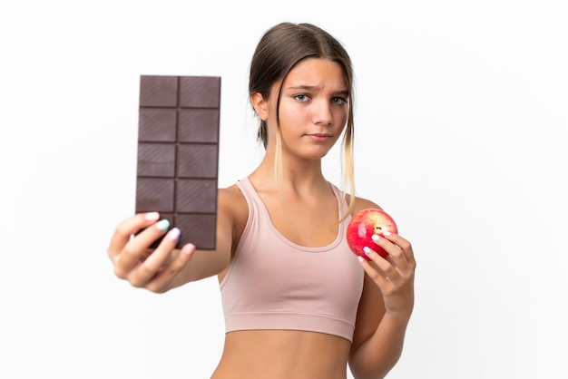 Petite fille caucasienne isolée sur fond blanc prenant une tablette de chocolat dans une main et une pomme dans l'autre