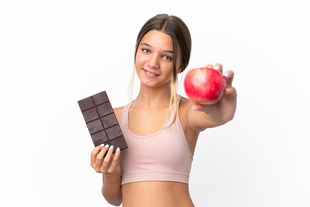 Petite fille caucasienne isolée sur fond blanc prenant une tablette de chocolat dans une main et une pomme dans l'autre