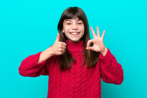 Petite fille caucasienne isolée sur fond bleu montrant le signe ok et le geste du pouce vers le haut