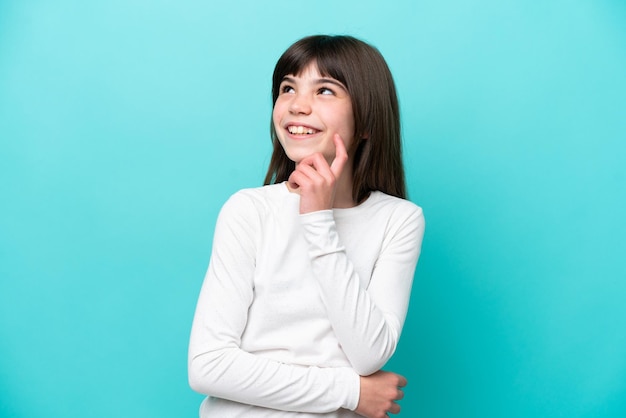 Petite fille caucasienne isolée sur fond bleu en pensant à une idée tout en levant les yeux