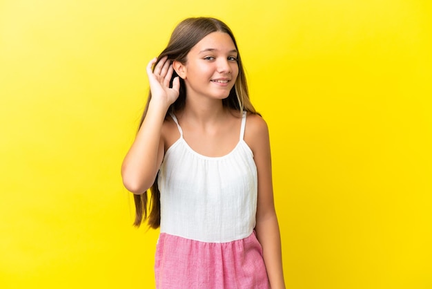 Petite fille caucasienne isolée sur fond jaune écoutant quelque chose en mettant la main sur l'oreille