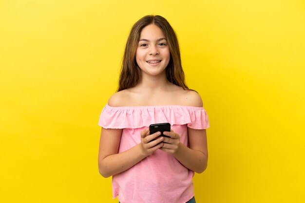 Petite fille caucasienne isolée sur fond jaune envoyant un message avec le mobile