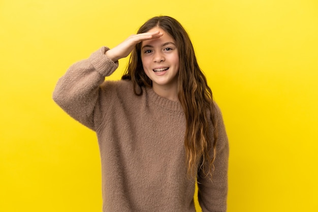 Petite fille caucasienne isolée sur fond jaune regardant loin avec la main pour regarder quelque chose