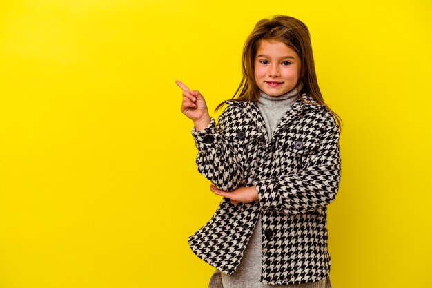 Petite fille caucasienne isolée sur fond jaune souriante pointant gaiement avec l'index loin.