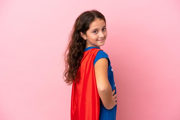 Petite fille caucasienne isolée sur fond rose en costume de super-héros et posant avec les bras à la hanche et souriant