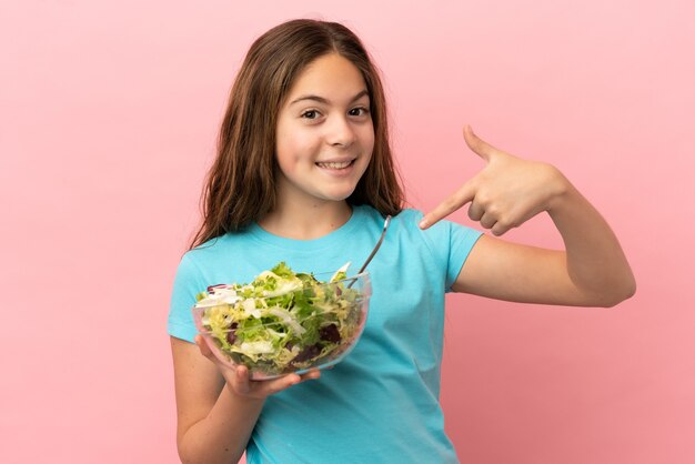 Petite fille caucasienne isolée sur fond rose avec une expression heureuse tout en pointant un bol de salade