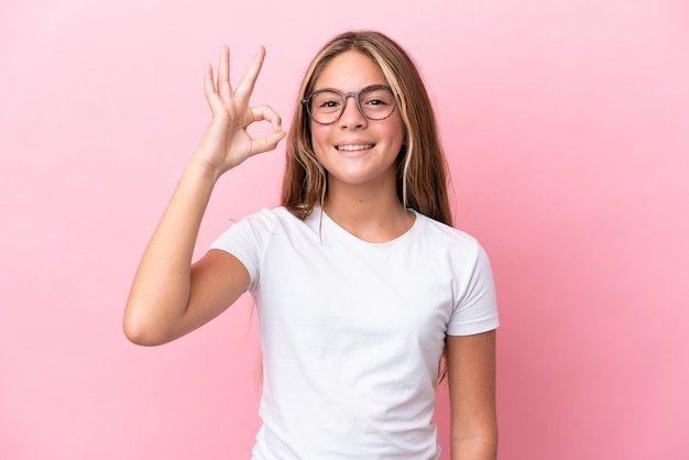 Petite fille caucasienne isolée sur fond rose avec des lunettes et faisant signe OK