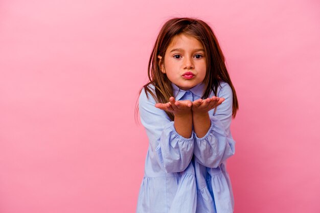 Petite fille caucasienne isolée sur fond rose pliant les lèvres et tenant les paumes pour envoyer un baiser aérien.