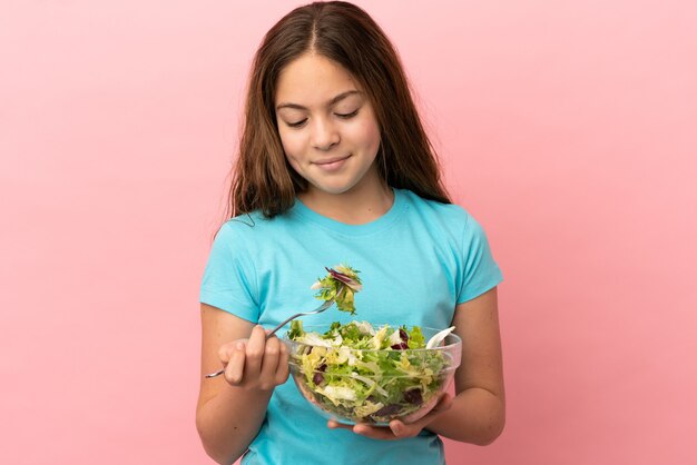 Petite fille caucasienne isolée sur fond rose tenant un bol de salade avec une expression heureuse