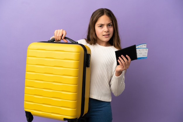 Petite fille caucasienne isolée sur fond violet malheureuse en vacances avec valise et passeport