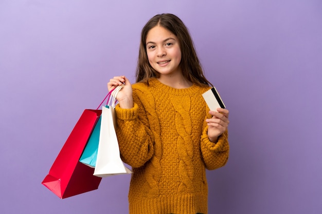 Petite fille caucasienne isolée sur fond violet tenant des sacs à provisions et une carte de crédit