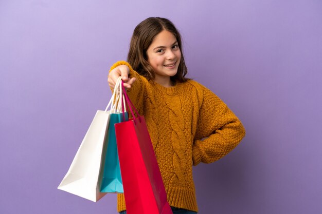 Petite fille caucasienne isolée sur fond violet tenant des sacs à provisions et les donnant à quelqu'un