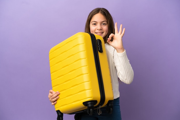 Petite fille caucasienne isolée sur fond violet en vacances avec valise de voyage et faisant signe OK