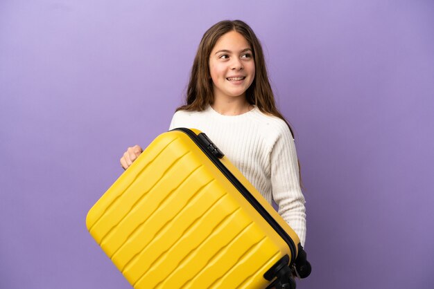 Petite fille caucasienne isolée sur fond violet en vacances avec valise de voyage