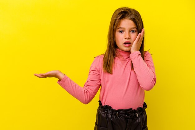 Petite fille caucasienne isolée sur jaune impressionné tenant un espace de copie sur la paume.