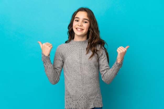 Petite fille caucasienne isolée sur un mur bleu avec un geste de pouce en l'air et souriant