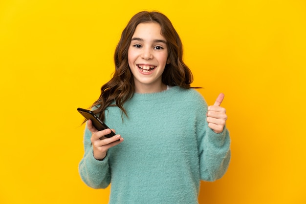 Petite fille caucasienne isolée sur un mur jaune à l'aide de téléphone mobile tout en faisant les pouces vers le haut