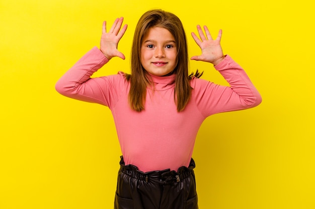 Petite fille caucasienne isolée sur un mur jaune recevant une agréable surprise, excitée et levant les mains.
