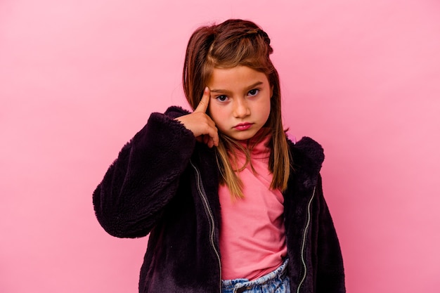 Petite fille caucasienne isolée sur le mur rose pointant le temple avec le doigt, pensant, concentré sur une tâche.