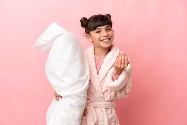 Petite fille caucasienne isolée sur rose en pyjama et invitant à venir avec la main