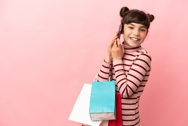 Petite fille caucasienne isolée sur rose tenant des sacs à provisions et appeler un ami avec son téléphone portable