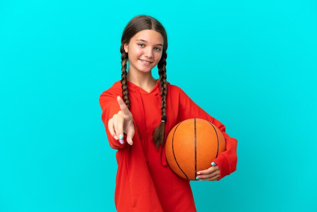 Petite Fille Caucasienne Jouant Au Basket-ball Isolée Sur Fond Bleu Montrant Et Levant Un Doigt