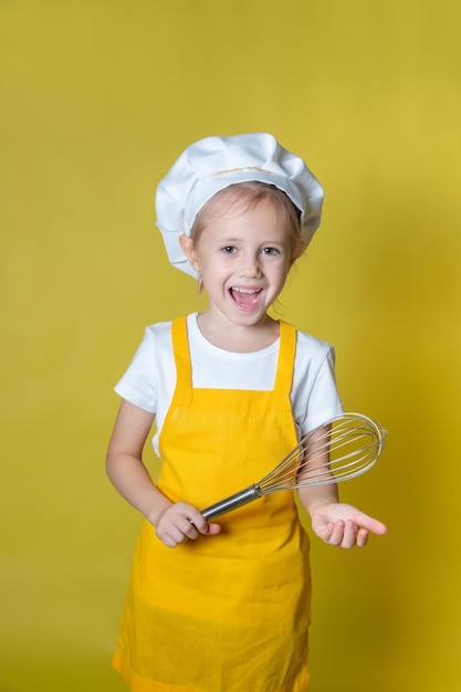 Petite fille caucasienne jouant au chef, fille en tablier et toque tenant un fouet pour fouetter la crème sur fond jaune