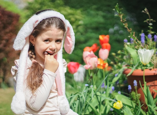 Une petite fille caucasienne avec des oreilles de lapin bandeau mange un œuf de pâques en chocolat en regardant la caméra