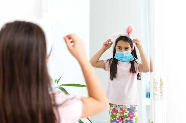 Petite fille caucasienne avec des oreilles de lapin et un masque médical sur son visage sur fond blanc. Notion de Pâques. Protégé contre le coronavirus