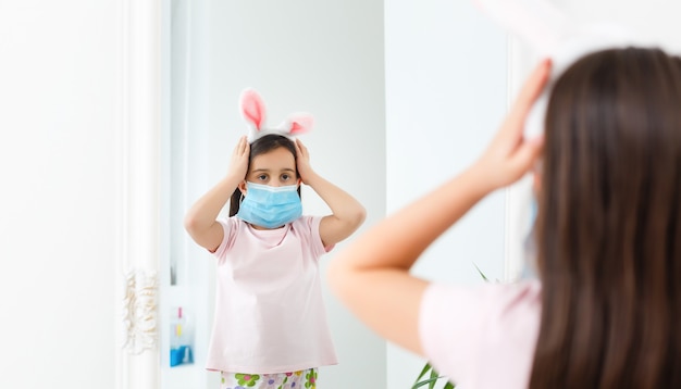 Petite fille caucasienne avec des oreilles de lapin et un masque médical sur son visage sur fond blanc. Notion de Pâques. Protégé contre le coronavirus