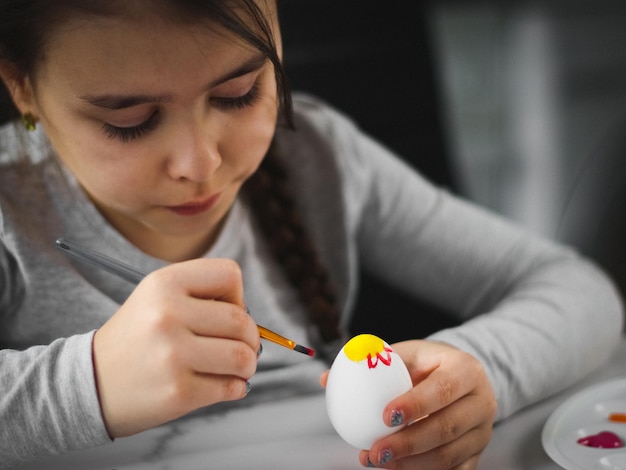 Une petite fille caucasienne peint avec enthousiasme un œuf de Pâques avec un pinceau de peinture acrylique