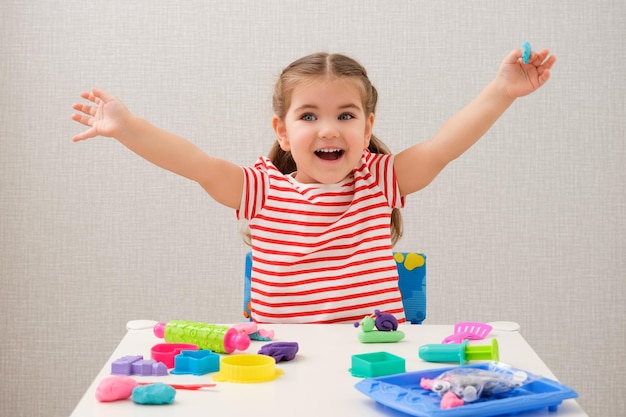 Petite fille caucasienne souriante blonde en T-shirt lumineux rayé joue avec de la pâte à modeler à la table blanche à la maison pâte à modeler