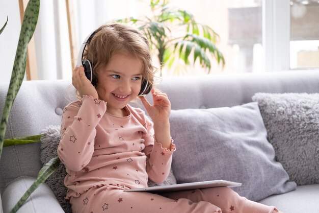 Petite fille caucasienne souriante et heureuse utilisant une tablette dans des écouteurs sans fil dans un canapé à la maison