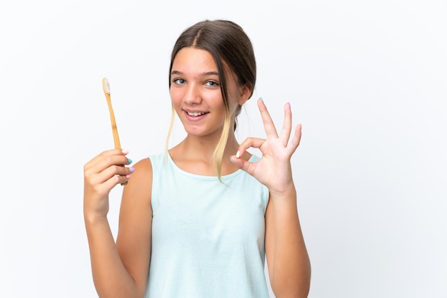 Petite fille caucasienne tenant une brosse à dents isolée sur fond blanc montrant un signe ok avec les doigts