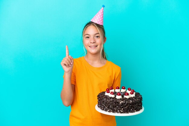 Petite fille caucasienne tenant un gâteau d'anniversaire isolé sur fond bleu montrant et levant un doigt en signe du meilleur