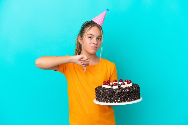 Petite fille caucasienne tenant un gâteau d'anniversaire isolé sur fond bleu montrant le pouce vers le bas avec une expression négative